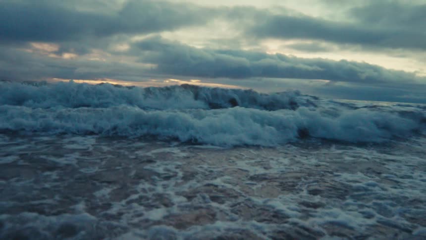 Intense slow-motion view from inside the water as powerful ocean waves crash over the camera under a stormy sky at dusk