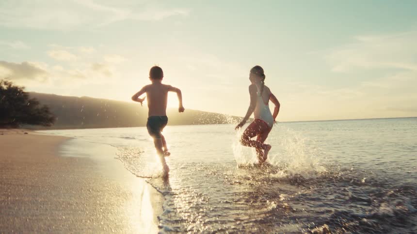 Two happy young kids run and splash in the shallow water along a sandy beach at sunset in slow motion