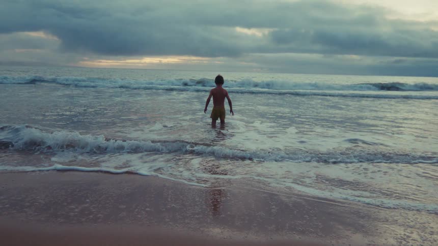 Slow motion shot of a young boy standing in the ocean waves as they wash ashore under a dramatic, cloudy sky