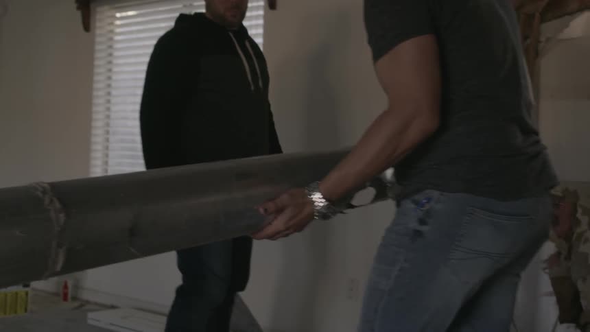Two men move a long pipe in a room