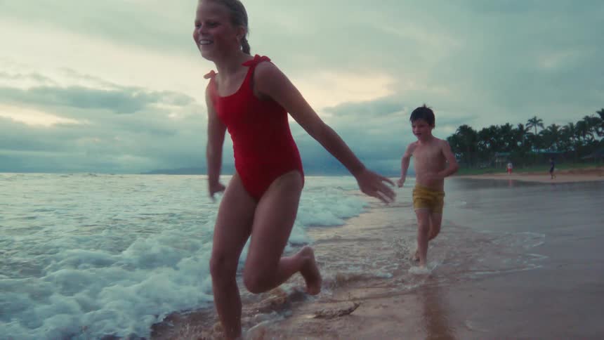 Three joyful children run and splash in the ocean waves along the sandy shore at dusk in slow motion