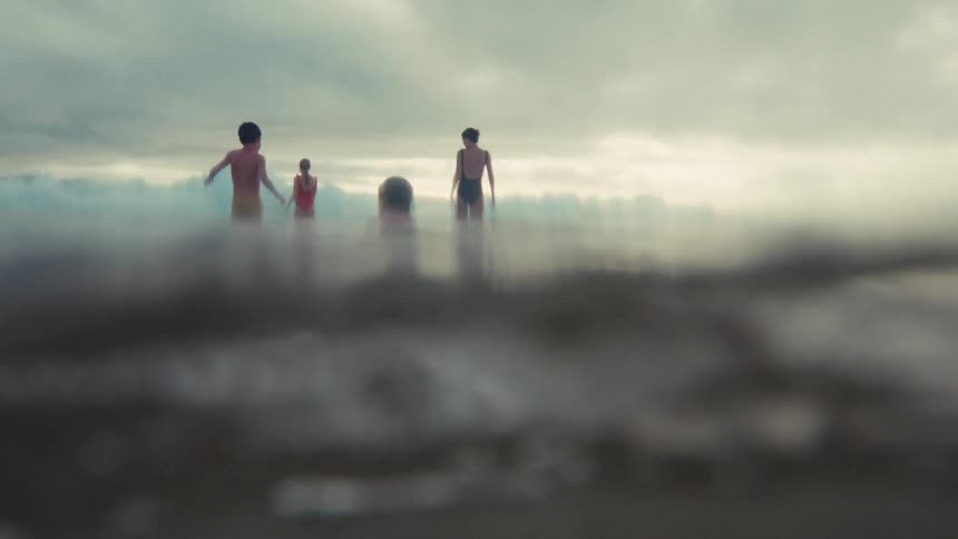 An immersive slow-motion view from the water's surface shows a family waiting for waves on a moody, overcast day