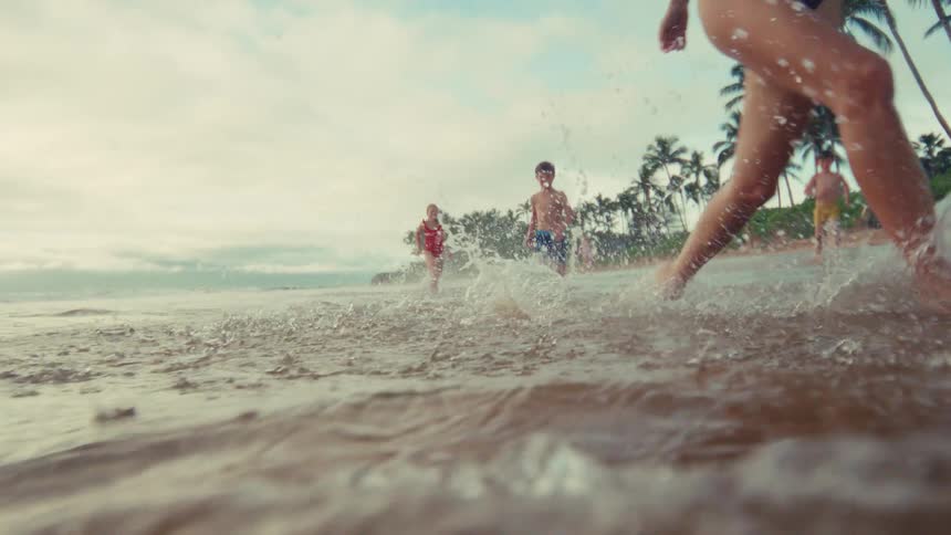 A slow motion, low angle shot of a woman and children joyfully running and splashing in the shallow waves of a tropical beach