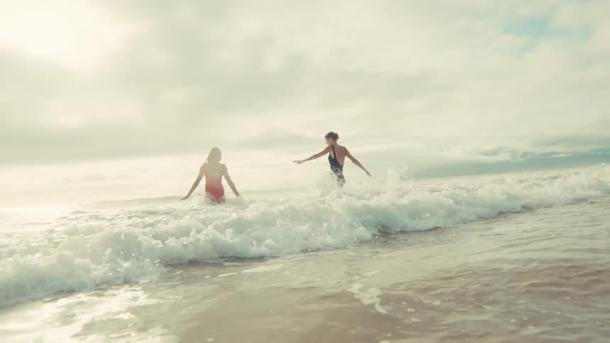 Two carefree women in swimsuits playfully run and jump in the shallow ocean waves in slow motion