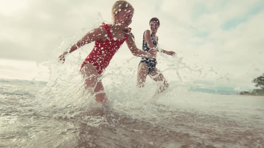 A smiling mother and daughter playfully splash and run through the shallow ocean water in slow motion