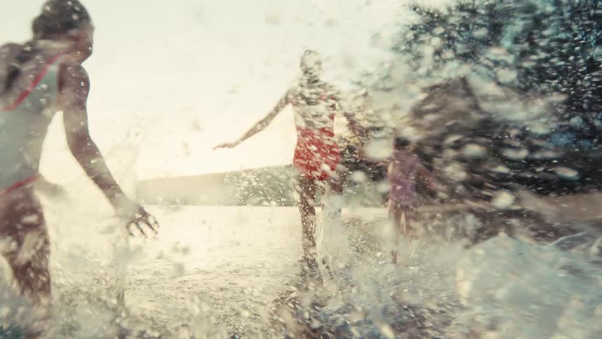 A mother and her two young daughters run hand-in-hand along a beautiful beach at sunset, splashing in the ocean waves in slow motion