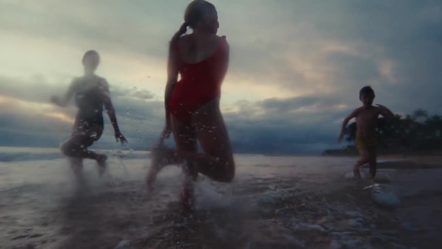 A family playfully runs through the shallow ocean water, splashing and jumping in dreamy slow motion against a dramatic twilight sky