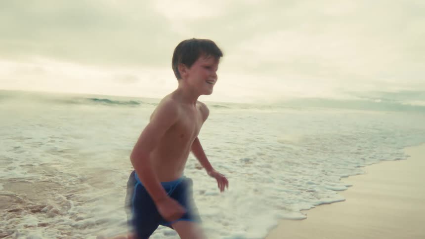 A happy young boy runs along the shoreline in slow motion as the ocean waves crash on the sandy beach