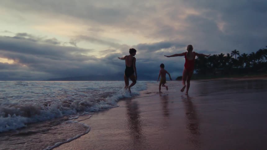 A happy family runs and plays together in the ocean waves on a tropical beach at sunset in slow motion