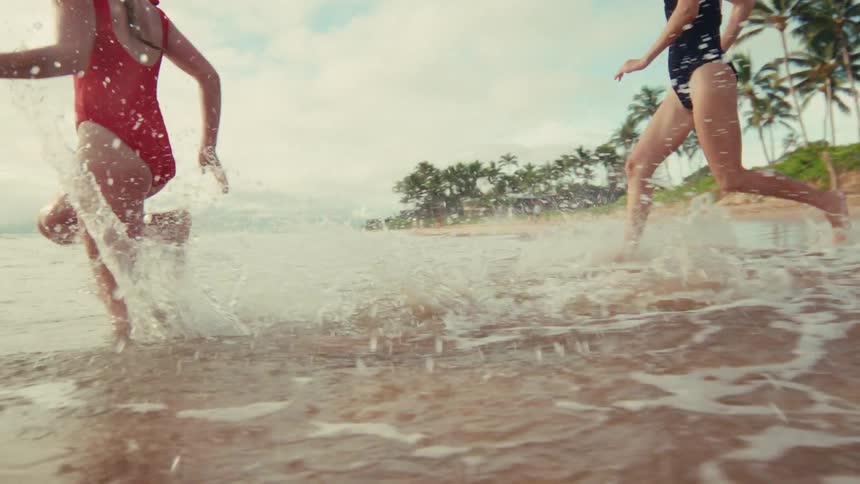 Two joyful young women in swimsuits run and splash into the ocean waves on a tropical beach in slow motion