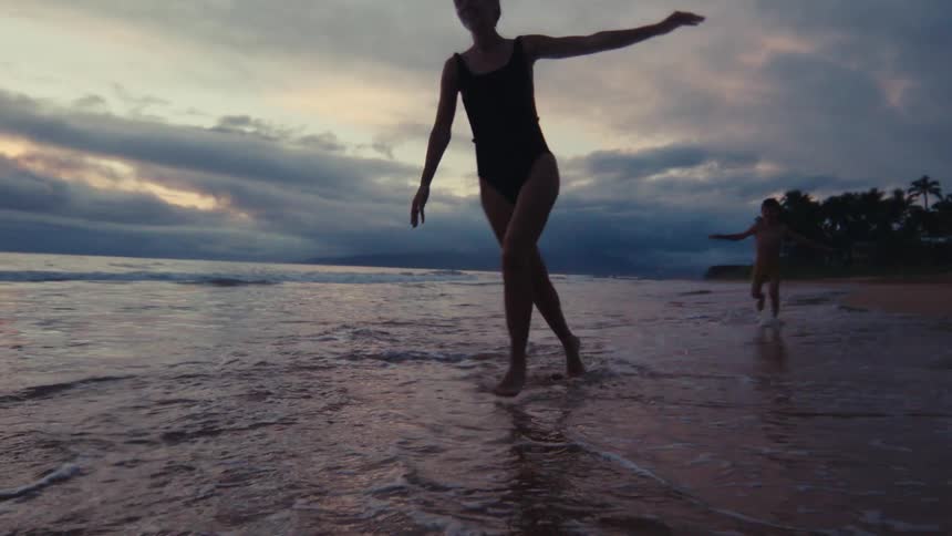 Women and a child joyfully run and splash through the shallow ocean waves on a tropical beach at dusk in slow motion