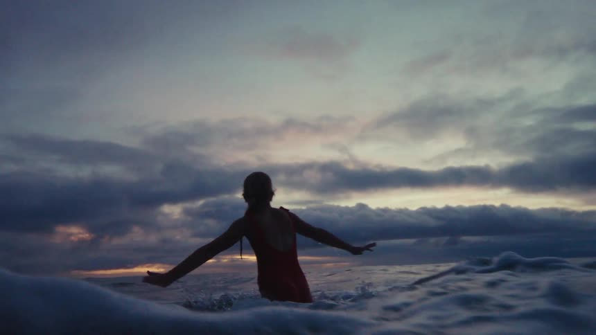 A young girl silhouetted in a red swimsuit joyfully jumps in the ocean waves during a beautiful, cloudy sunset