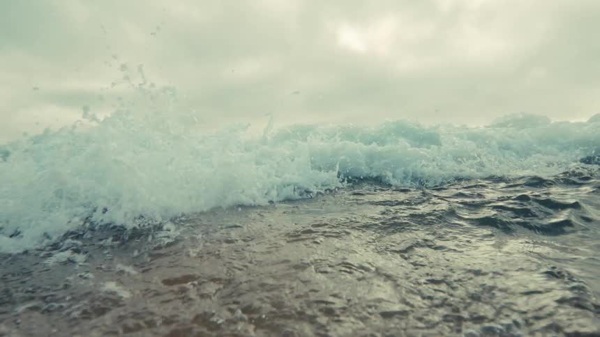A dramatic slow-motion shot from water level as an ocean wave builds and crashes over the camera under a cloudy sky