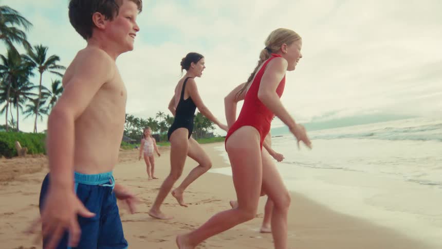 A joyful family with young children excitedly run along the sandy shoreline in slow motion on a beautiful tropical beach