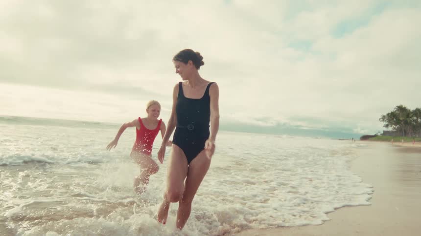 A happy mother and her young daughter run and splash in the gentle ocean waves along a sandy tropical beach in slow motion