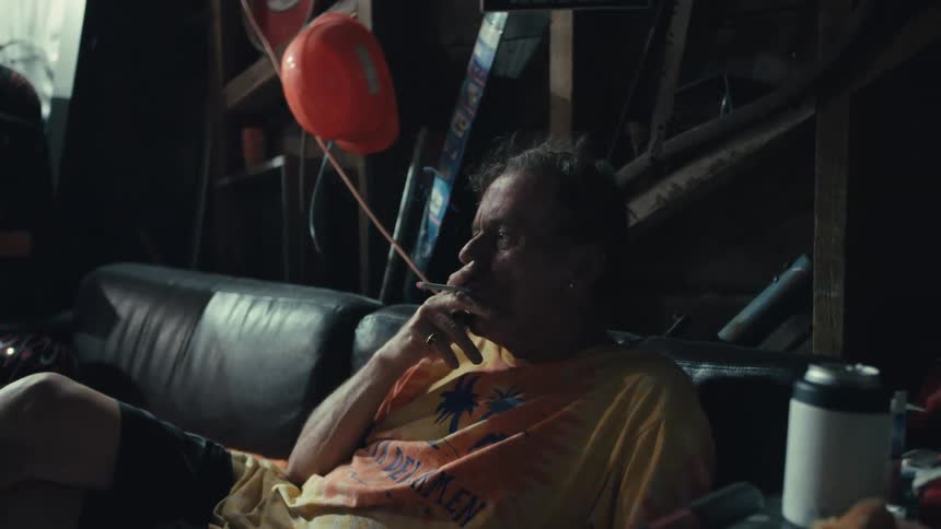 An older man in a tie-dye shirt smokes a cigarette in slow motion while sitting on a couch in a cluttered garage