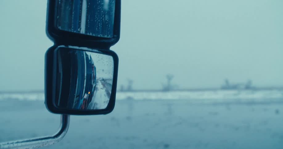 Close-up of a semi-truck's side view mirror reflecting the snowy highway on a cold, overcast winter day