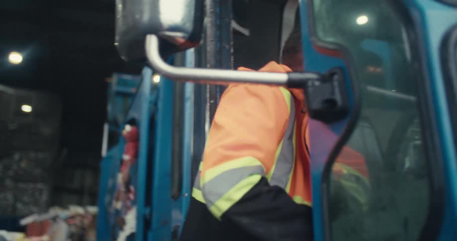A male sanitation worker gets into his garbage truck inside a large, dimly lit recycling plant