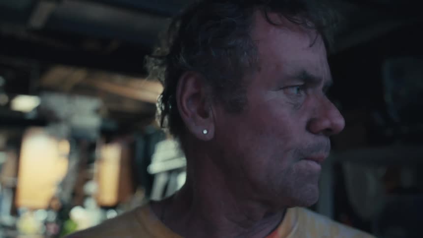 A rugged, middle-aged man with a dirty face thoughtfully smokes a cigarette in a dimly lit workshop
