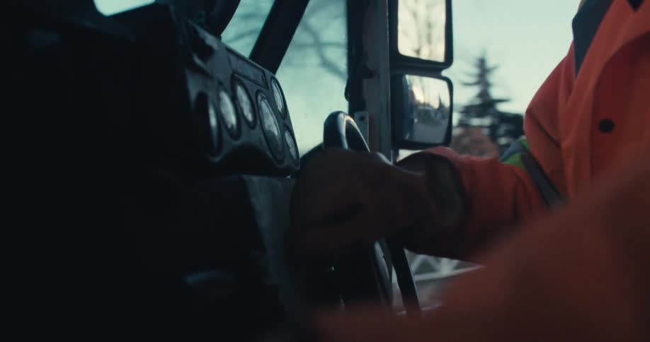 An over-the-shoulder view from inside a truck cab shows a worker in an orange jacket driving down a road at dusk