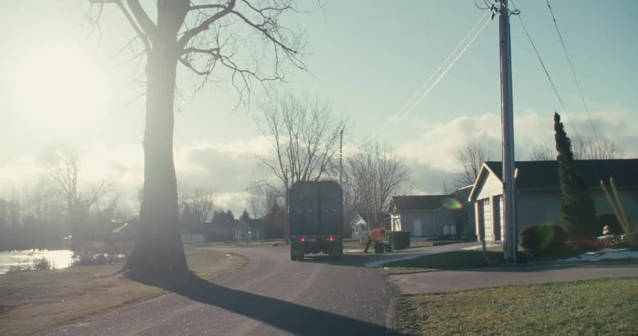 A sanitation worker empties a curbside recycling bin into a garbage truck on a sunny, hazy morning in a quiet neighborhood