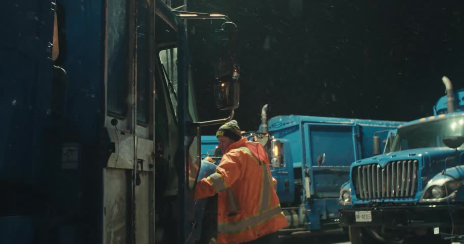 A garbage truck driver in a high-visibility jacket exits his cab and inspects the truck's tire on a snowy winter night