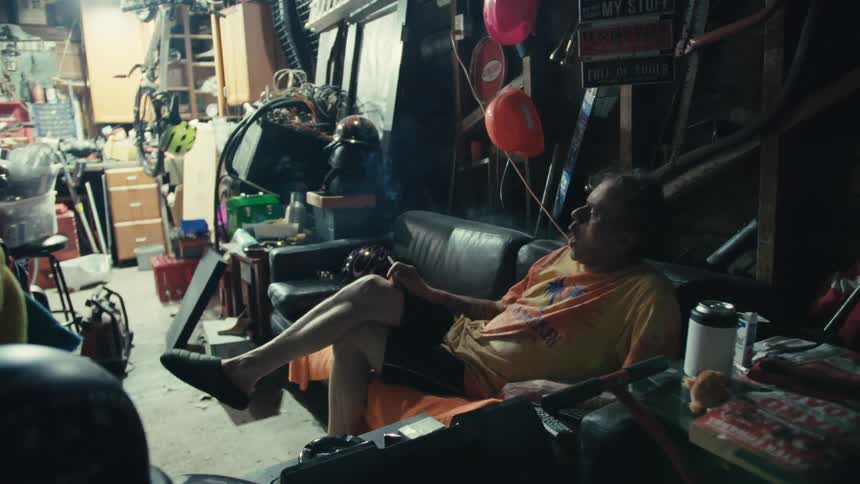 An older man relaxes on a leather couch and smokes a cigarette in his cluttered garage workshop