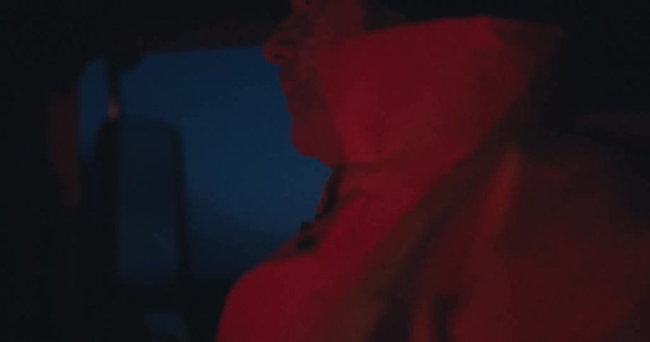 Moody close-up of a man smoking a cigarette inside his vehicle at night, his face cast in a dramatic red glow