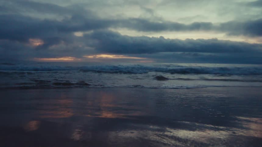 Ocean waves gently wash ashore in slow motion under a moody, cloudy sky during twilight