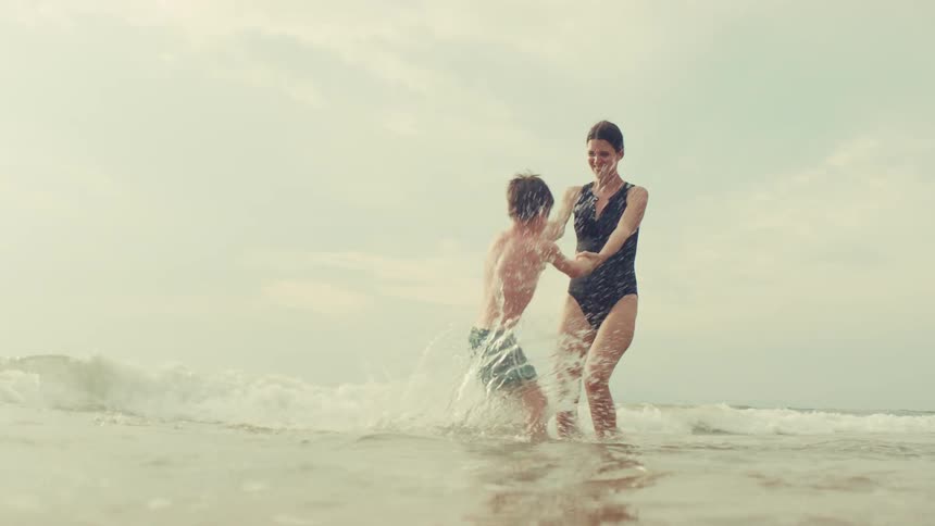 A mother and her young son playfully spin and splash in the ocean waves, holding hands and smiling in slow motion