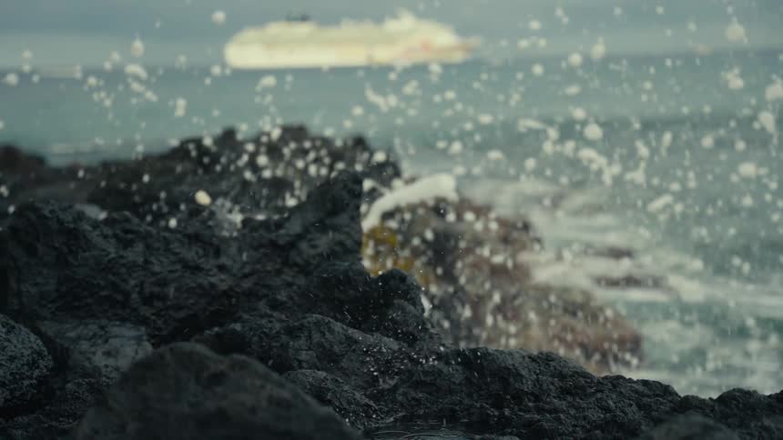 Ocean waves crash in slow motion against a dark, rocky shore with a cruise ship visible in the distance