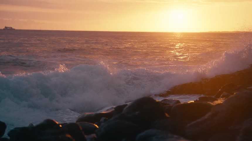 A slow-motion shot of powerful ocean waves crashing against a rocky shoreline during a vibrant golden sunset