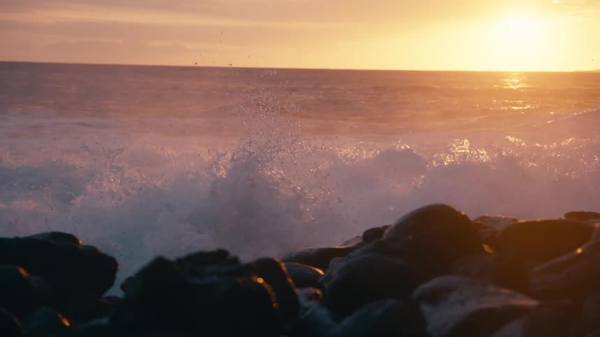 A powerful ocean wave crashes against a rocky shoreline in dramatic slow motion as the golden sun sets on the horizon