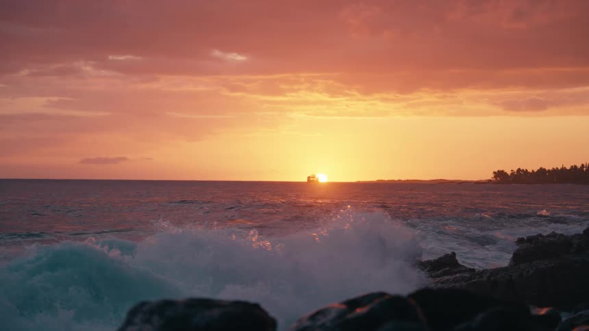 A slow-motion view shows powerful ocean waves crashing over rocks during a vibrant sunset with a distant boat on the horizon