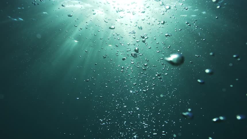 Slow motion shot of air bubbles rising through the deep blue ocean as sun rays beam down from the surface