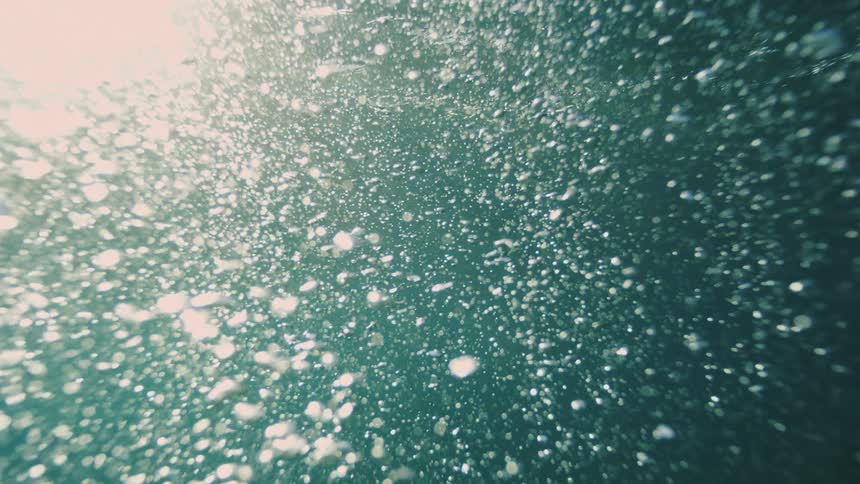 Sunlit underwater scene shows thousands of tiny bubbles rising through turquoise water