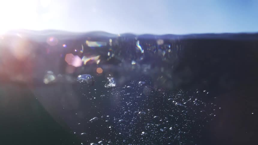 Graceful air bubbles rise in slow motion through dark water, breaking the surface to reveal a sunlit sky