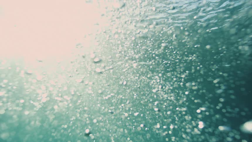 An abstract underwater perspective shows thousands of tiny bubbles rising towards the bright sunlit surface in slow motion