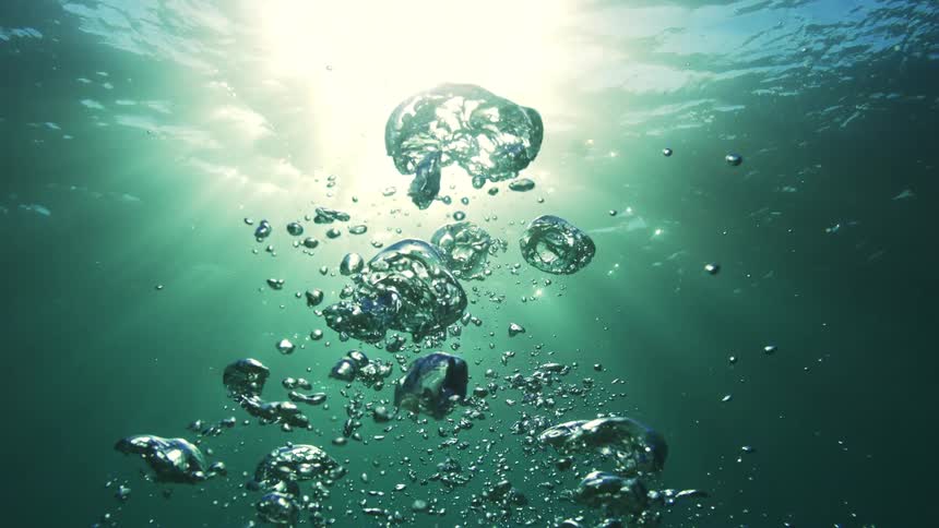 Super slow motion shot of air bubbles rising through the teal ocean water towards the bright sun