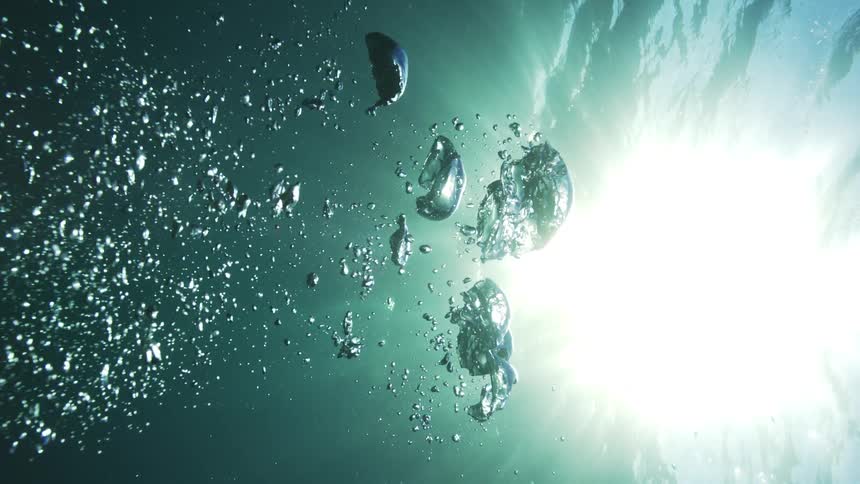 Slow motion bubbles rise through the teal water towards the bright sunlit surface