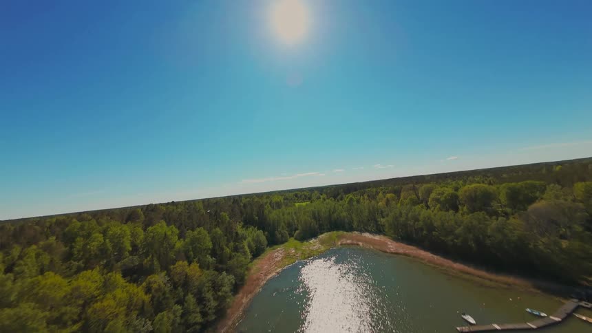 An aerial FPV drone soars over a sunlit lake and dense green forest, tilting up to reveal the brilliant sun in a clear blue sky