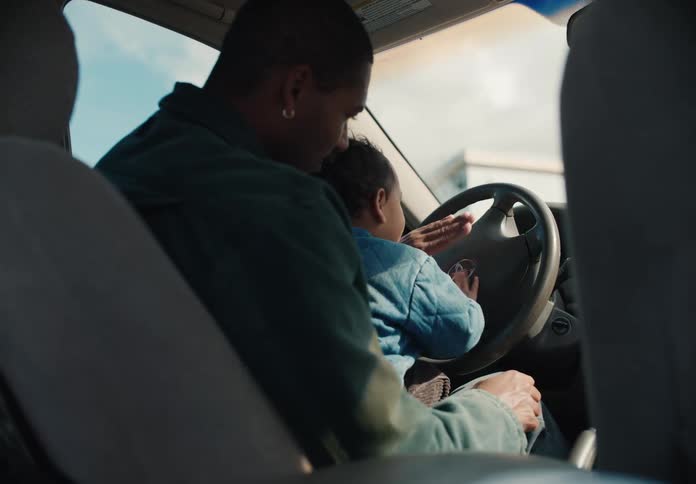 A smiling father holds his toddler on his lap in the driver's seat as the child playfully honks the car horn