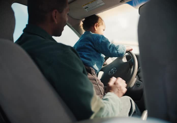 A loving father holds his toddler on his lap in the driver's seat, letting them playfully turn the steering wheel