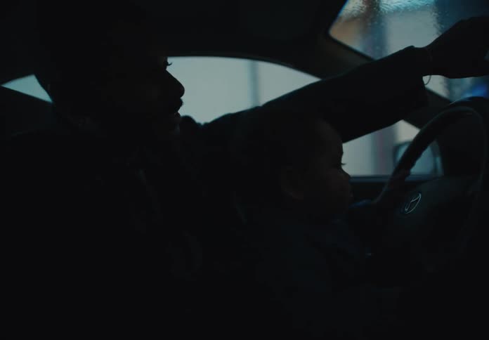 A father holds his toddler on his lap in the driver's seat of a car at night, pointing out the window as the child watches