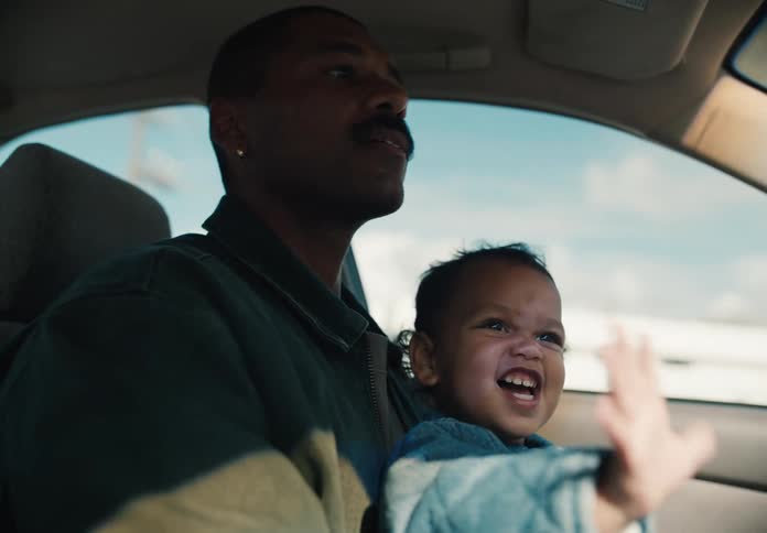 A cute toddler smiles and waves happily while sitting on his father's lap as he drives a car on a sunny day