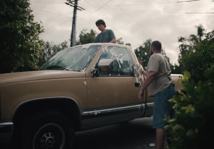 A father and his teenage son work together to wash their old pickup truck in the driveway on an overcast day