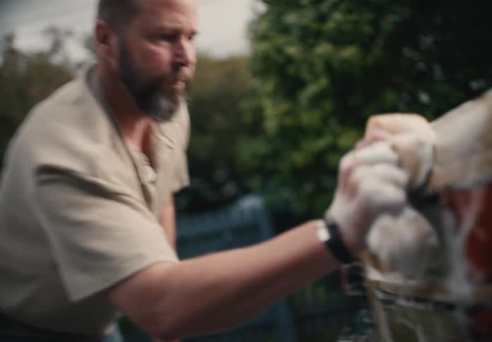 A bearded man washes the taillight of his vintage car with a soapy sponge in slow motion