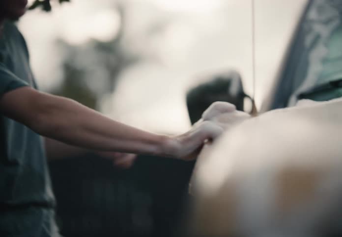 A teenage boy smiles as he washes the hood of a sudsy car with a sponge in the driveway
