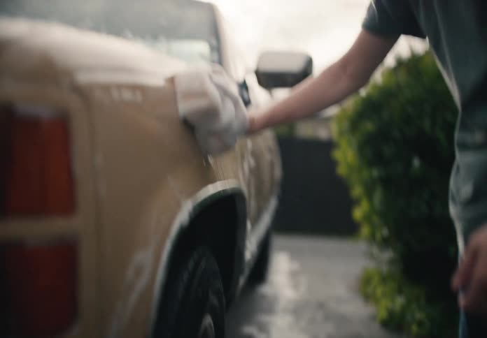 A young man carefully washes his vintage tan pickup truck with a soapy sponge in the driveway of his home