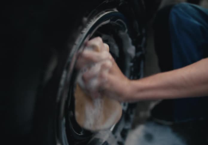 A focused young man kneels down and scrubs the soapy wheel of a car with a sponge